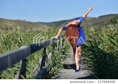Woman with red hair in blue dress standing on wooden footbridge in a reed field Woman with red hair in blue dress standing on wooden footbridge in a reed field 137898400