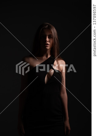 Young woman creating a sense of mystery and elegance, wearing a black evening dress with a cutout design, her face partially illuminated in a low-key studio setting Young woman creating a sense of mystery and elegance, wearing a black evening dress with a cutout design, her face partially illuminated in a low-key studio setting 137898597