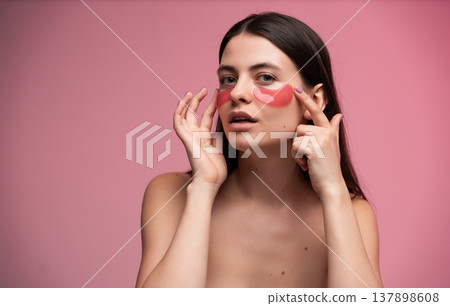 Young woman applying red hydrogel eye patches for under-eye skincare, receiving beauty treatment for tired eyes, rejuvenating and moisturizing facial skin on pink background Young woman applying red hydrogel eye patches for under-eye skincare, receiving beauty treatment for tired eyes, rejuvenating and moisturizing facial skin on pink background 137898608