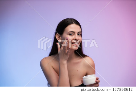 Young woman applying face cream, moisturizing her skin for beauty and self-care, promoting healthy glow and daily skincare routine against a vibrant gradient background Young woman applying face cream, moisturizing her skin for beauty and self-care, promoting healthy glow and daily skincare routine against a vibrant gradient background 137898612