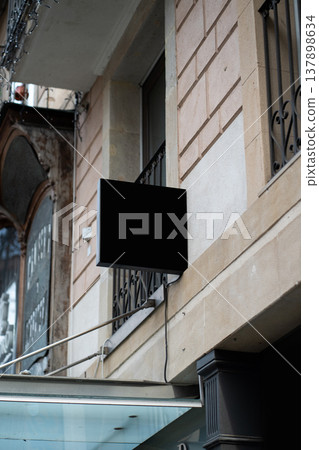 Blank black street sign mock-up on building facade, ready for branding and advertising copy space, hanging above a shop awning on an old city street with classic architecture 137898634