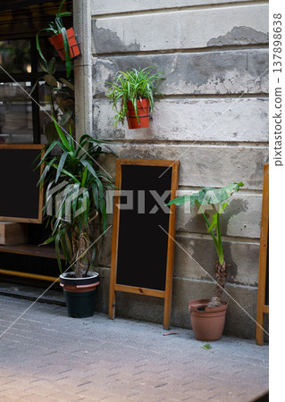Sidewalk cafe or restaurant entrance with empty menu chalkboards, surrounded by diverse potted plants and lush urban greenery along a textured concrete wall, waiting for customization Sidewalk cafe or restaurant entrance with empty menu chalkboards, surrounded by diverse potted plants and lush urban greenery along a textured concrete wall, waiting for customization 137898638