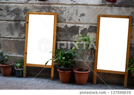 Two blank wooden sandwich boards offering advertising mockups and branding space, standing on old street pavement near a grungy wall with potted plants Two blank wooden sandwich boards offering advertising mockups and branding space, standing on old street pavement near a grungy wall with potted plants 137898639