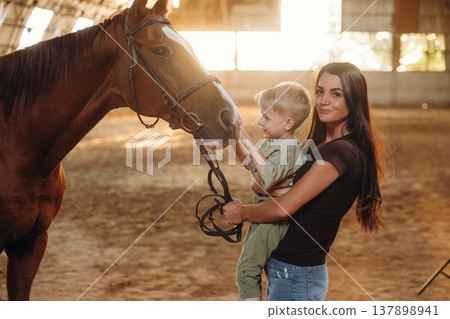 Curious kid is touching the animal. Mother and little son are with horse in hangar 137898941