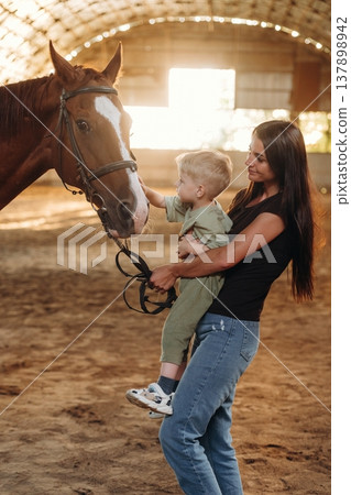 Curious kid is touching the animal. Mother and little son are with horse in hangar 137898942