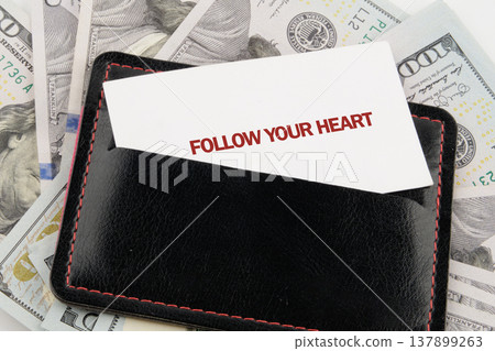 A stark white card urging FOLLOW YOUR HEART emerges from a sleek black wallet, resting upon a backdrop of US Dollar hundred dollar bills 137899263