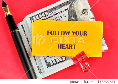 A poignant yellow card with a guiding phrase, surrounded by plentiful United States currency and an elegant writing instrument, all on a striking red field A poignant yellow card with a guiding phrase, surrounded by plentiful United States currency and an elegant writing instrument, all on a striking red field 137899284