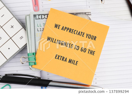 A vibrant orange appreciation note recognizing hard work rests on a stack of United States currency, accompanied by office supplies on a planner surface A vibrant orange appreciation note recognizing hard work rests on a stack of United States currency, accompanied by office supplies on a planner surface 137899294