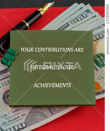 A message on a green card emphasizes the critical role of contributions in fostering achievements, laid upon US dollar bills and a sophisticated fountain pen 137899304