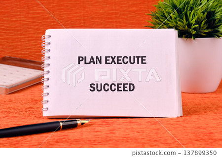 A bright spiral notebook showcasing the words PLAN EXECUTE SUCCEED sits on an orange desk with a pen and a small green plant A bright spiral notebook showcasing the words PLAN EXECUTE SUCCEED sits on an orange desk with a pen and a small green plant 137899350