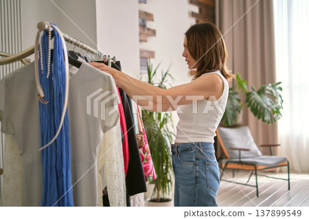 stylist in a white outfit examines various clothing items on a rack, showcasing stylist in a white outfit examines various clothing items on a rack, showcasing 137899549