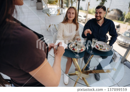 The waiter stands and takes the order. Couple enjoying date in the outdoor restaurant The waiter stands and takes the order. Couple enjoying date in the outdoor restaurant 137899559