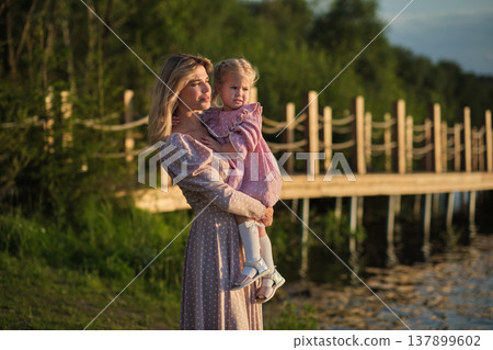 Contemplative mother holds child near lake at dusk. A tender moment evoking thoughts of protection and future dreams. 137899602