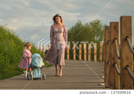 Strolling on a wooden bridge, a mother and daughter enjoy an evening walk. The scene echoes everyday family leisure. Strolling on a wooden bridge, a mother and daughter enjoy an evening walk. The scene echoes everyday family leisure. 137899616