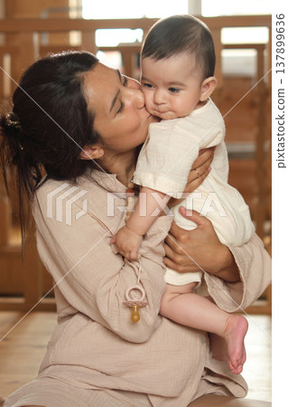 Family Foundations: Witness a mother's pure joy as she snuggles her one-year-old, framed by the wooden architecture of their snug home Family Foundations: Witness a mother's pure joy as she snuggles her one-year-old, framed by the wooden architecture of their snug home 137899636
