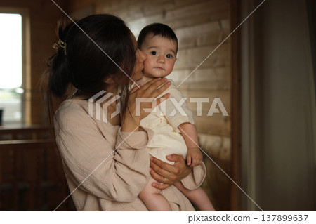 Nurturing Love: Inside a charming timber house, a mother finds her happy place, holding her baby girl tight and stealing sweet kisses. 137899637