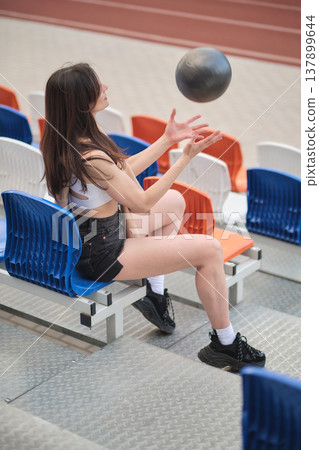 Woman in casual sportswear, throwing a black ball in the air while seated, showcasing balance and control. Concept: Demonstrating the role of simple, engaging activities in promoting wellness and Woman in casual sportswear, throwing a black ball in the air while seated, showcasing balance and control. Concept: Demonstrating the role of simple, engaging activities in promoting wellness and 137899644