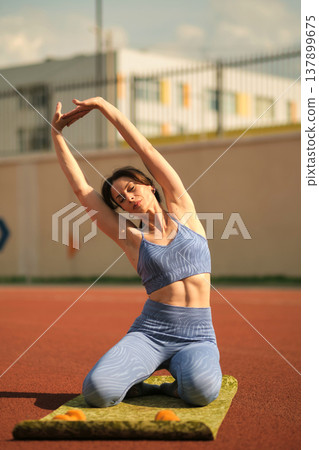 woman in blue sportswear performs a side stretch on a mat under the sunny sky. This exercise emphasizes the importance of flexibility and relaxation in a balanced fitness routine. woman in blue sportswear performs a side stretch on a mat under the sunny sky. This exercise emphasizes the importance of flexibility and relaxation in a balanced fitness routine. 137899675