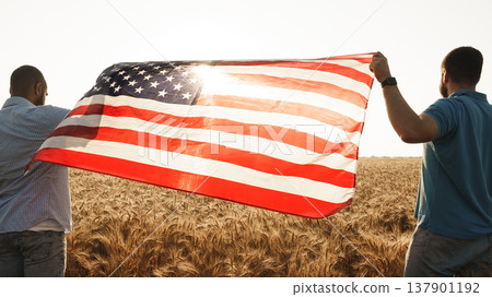 Two men hold an American flag in a wheat field during sunset hours Two men hold an American flag in a wheat field during sunset hours 137901192