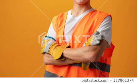 Worker poses with arms crossed in safety gear against orange background Worker poses with arms crossed in safety gear against orange background 137901374