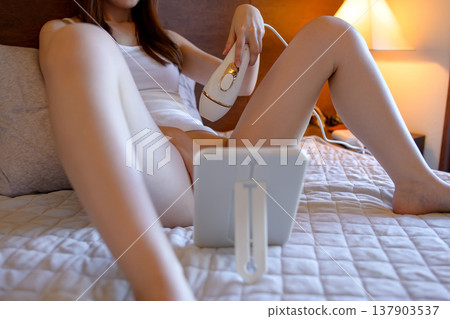 一位年輕女子正在使用家用除毛儀自行進行比基尼部位除毛;斜角視角。 一位年輕女子正在使用家用除毛儀自行進行比基尼部位除毛;斜角視角。 137903537
