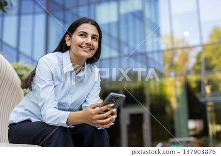 Female professional sitting on a modern bench outdoors, holding a smartphone and smiling while looking up, representing communication, innovative thinking, and career development Female professional sitting on a modern bench outdoors, holding a smartphone and smiling while looking up, representing communication, innovative thinking, and career development 137903876