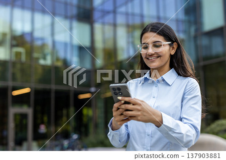 Happy young indian businesswoman reading or sending messages on her cellphone while standing outdoors in front of a modern glass office building, representing mobile communication and technology 137903881