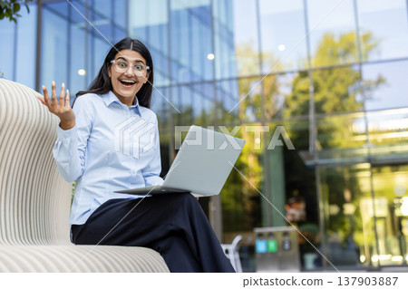 Excited woman in glasses celebrating good news or personal achievement while working on a laptop outside a modern office building, representing career success and entrepreneurship Excited woman in glasses celebrating good news or personal achievement while working on a laptop outside a modern office building, representing career success and entrepreneurship 137903887