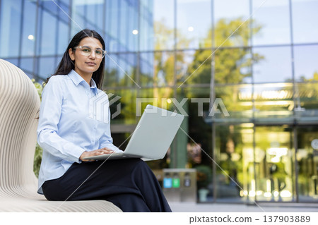 Young woman in formal wear sitting on a modern bench, confidently using her laptop outdoors, working remotely with a modern glass office building in the background Young woman in formal wear sitting on a modern bench, confidently using her laptop outdoors, working remotely with a modern glass office building in the background 137903889