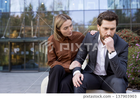 Woman showing empathy and support, placing hands on a stressed man's shoulder and arm, offering consolation during a difficult work situation outside a modern office 137903903