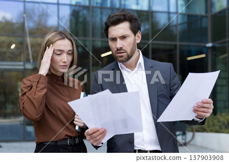 Two business professionals outside an office building examine financial documents with worried, frustrated expressions as they grapple with an urgent report, possible errors and risk Two business professionals outside an office building examine financial documents with worried, frustrated expressions as they grapple with an urgent report, possible errors and risk 137903908