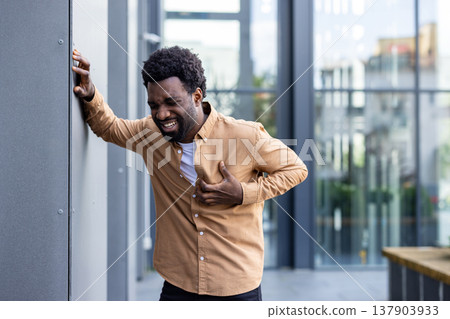Adult individual experiencing sharp pains in their chest area, grasping their shirt while leaning against a wall outdoors, indicating discomfort and potential health suffering 137903933