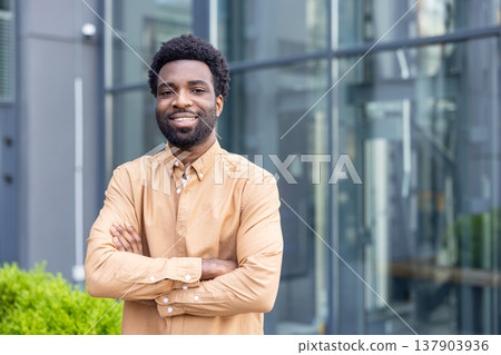 Young african american professional standing outdoors with arms crossed, smiling confidently in front of glass-fronted urban architecture, conveying ambition and success 137903936