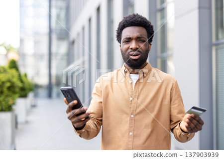 African american man feeling confused and frustrated while trying to make an online payment with his credit card and smartphone, encountering a problem outdoors African american man feeling confused and frustrated while trying to make an online payment with his credit card and smartphone, encountering a problem outdoors 137903939