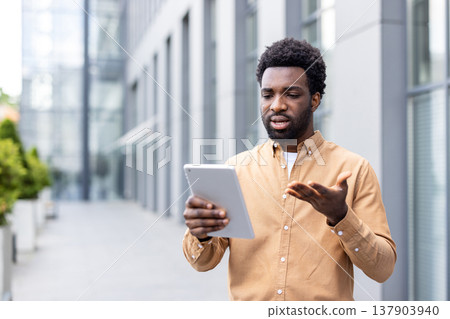 Young professional man standing outdoors, expressing frustration and confusion while looking at and gesturing towards a digital tablet, possibly facing tech issues Young professional man standing outdoors, expressing frustration and confusion while looking at and gesturing towards a digital tablet, possibly facing tech issues 137903940