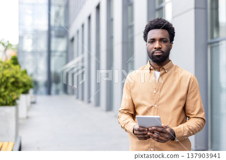 African executive standing outside a modern office building, holding a digital tablet with both hands, representing business professionalism, mobile technology, and urban connectivity African executive standing outside a modern office building, holding a digital tablet with both hands, representing business professionalism, mobile technology, and urban connectivity 137903941