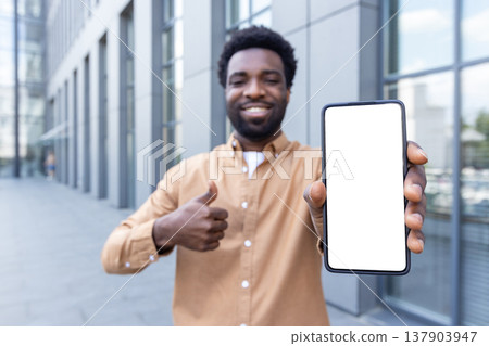 Smiling man in casual business attire outdoors, presenting a smartphone with a white blank screen for mockups while giving a thumbs up, confident and promotional in an urban setting 137903947