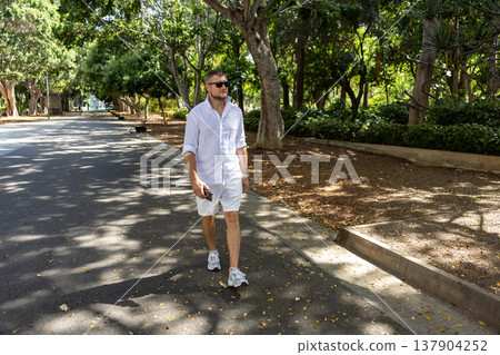 Casual Man Walking In Green Urban Park On Sunny Summer Day With Smartphone Casual Man Walking In Green Urban Park On Sunny Summer Day With Smartphone 137904252
