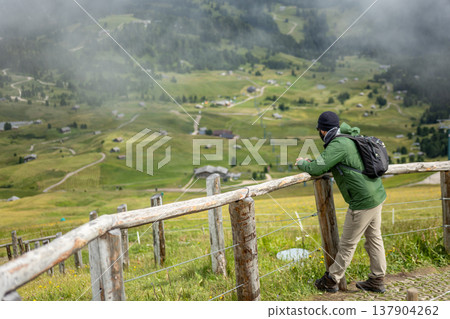 A traveler with a backpack stands at a wooden fence, admiring a wide alpine valley below. A traveler with a backpack stands at a wooden fence, admiring a wide alpine valley below. 137904262