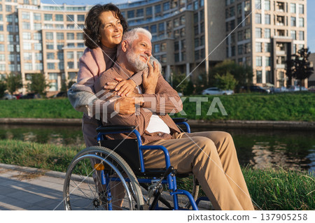Old man in wheelchair walking with caregiver senior woman on road in park. Elderly family couple woman supporting embracing paralyzed man in chair for people with disability outdoor. Rehabilitation Old man in wheelchair walking with caregiver senior woman on road in park. Elderly family couple woman supporting embracing paralyzed man in chair for people with disability outdoor. Rehabilitation 137905258