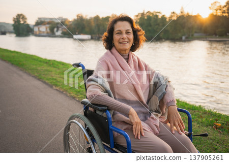 Happy senior woman in wheelchair smiling enjoying freedom walking on road in city park outdoor. Old cheerful positive woman in chair for people with disability. Disability disease rehabilitation Happy senior woman in wheelchair smiling enjoying freedom walking on road in city park outdoor. Old cheerful positive woman in chair for people with disability. Disability disease rehabilitation 137905261