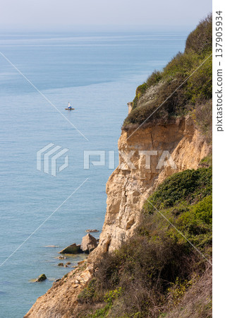 Chalk cliffs overlooking the sea with a small sailboat on the calm blue water 137905934