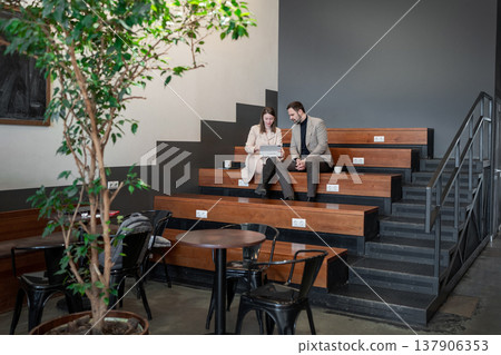 Partners going over notes together on wooden office steps in plant-filled workspace. Notes, plant-filled workspace, office, teamwork, partners. 137906353