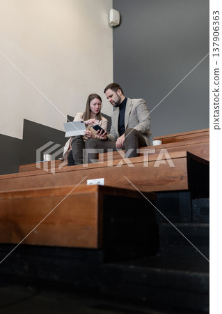 Partners collaborating on project on wooden office steps, communicating in relaxed workspace. Project, relaxed workspace, partners, collaboration, office. Partners collaborating on project on wooden office steps, communicating in relaxed workspace. Project, relaxed workspace, partners, collaboration, office. 137906363