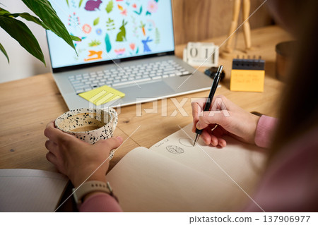 Woman holds pen and draws in notebook with laptop and coffee mug in home workspace 137906977