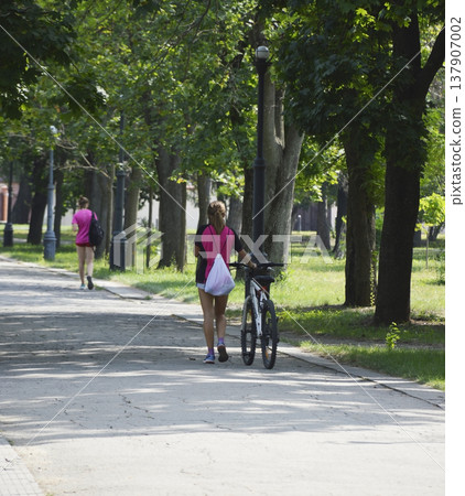 a beautiful girl walks along a path in the park and rolls a bicycle next to her 137907002