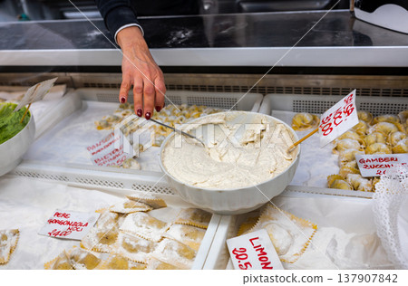 Genoa, liguria, italy. May 18, 2024. Woman's hand serving creamy filling next to trays of traditional italian pasta, ravioli, and pansoti at genoa oriental market 137907842