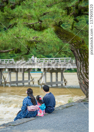 一家人穿著浴衣(夏季和服),背對鏡頭,坐在嵐山渡輪附近的河邊。日本京都府京都市 一家人穿著浴衣(夏季和服),背對鏡頭,坐在嵐山渡輪附近的河邊。日本京都府京都市 137909520