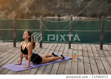 Beautiful Woman Practicing Cobra Yoga Pose On Mat At Lake. Spine Stretch And Flexibility Training Concept Beautiful Woman Practicing Cobra Yoga Pose On Mat At Lake. Spine Stretch And Flexibility Training Concept 137909721