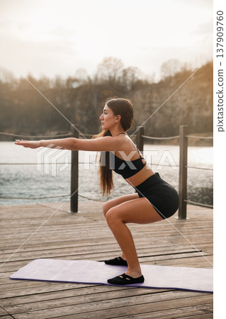 Young Fit Woman Doing Squat With Arms Extended Forward At Lake. Coordination And Strength Training Concept Young Fit Woman Doing Squat With Arms Extended Forward At Lake. Coordination And Strength Training Concept 137909760